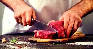 Butcher slicing fresh raw beef steak on wooden cutting board with herbs